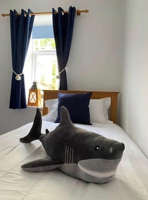 Una cama con un gran almohadón con forma de tiburón, cabecera de madera y ventana con cortinas.