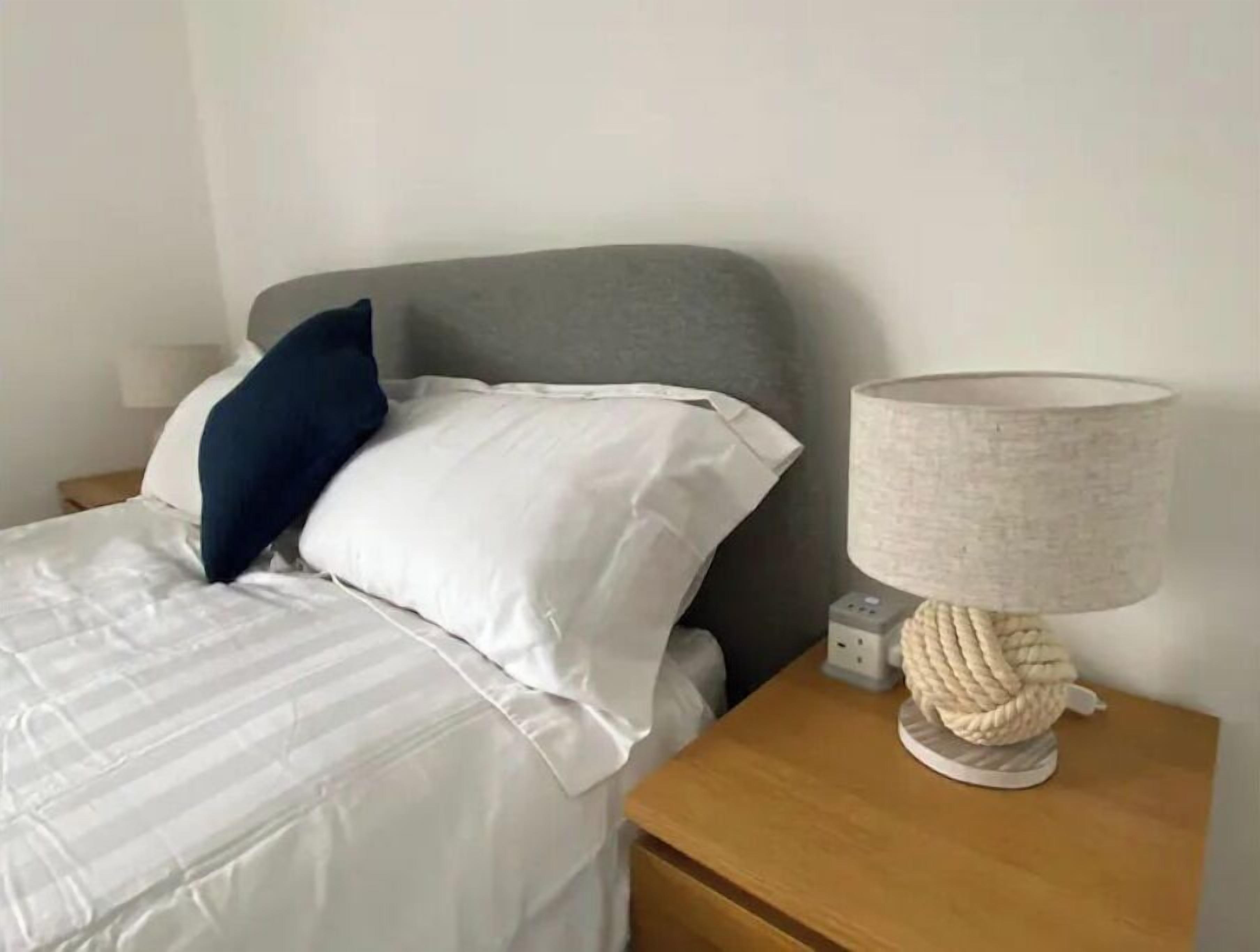 Una cama bien hecha con ropa blanca y una almohada azul, cabecera gris y mesita de noche con lámpara e interruptorio.