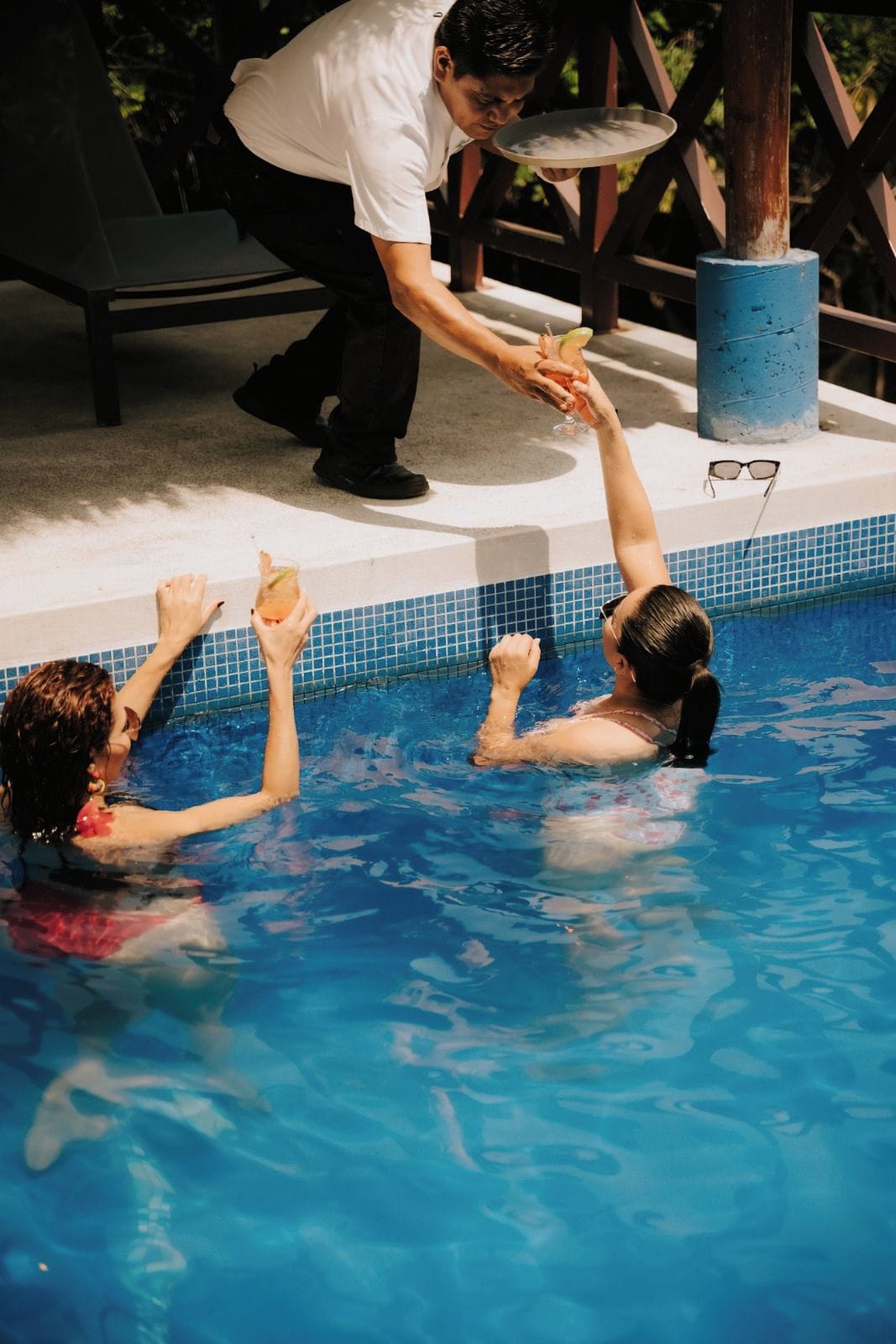 A poolside scene with a person serving drinks to two individuals in the water.