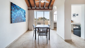 A dining area with a table and chairs, a ceiling fan, and a large window with a view of buildings. - Villas Coral Huatulco (Santa María Huatulco)