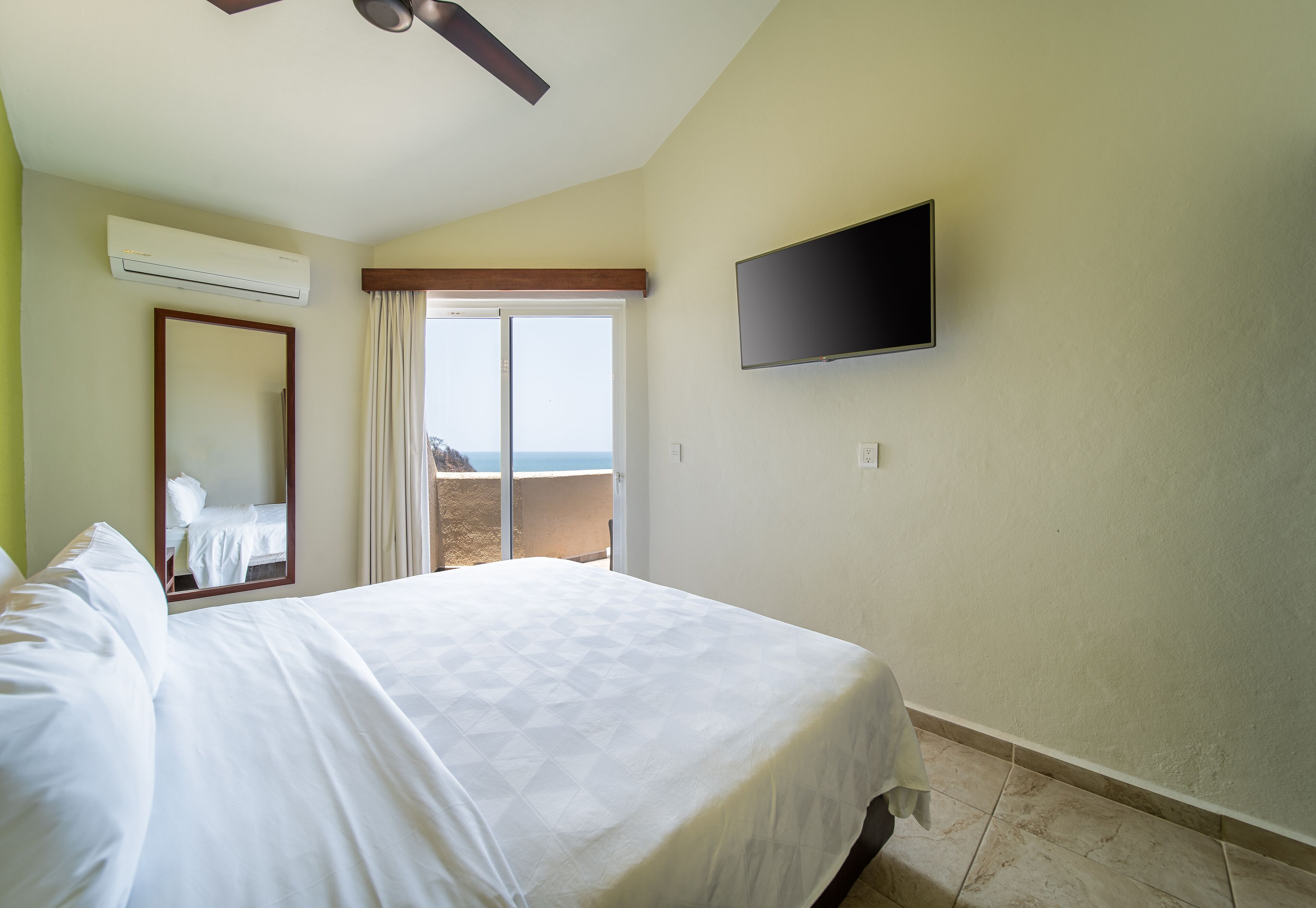 A hotel room with a bed, a flat-screen TV mounted on the wall, an air conditioning unit, a large window with a view of the beach, and a ceiling fan.