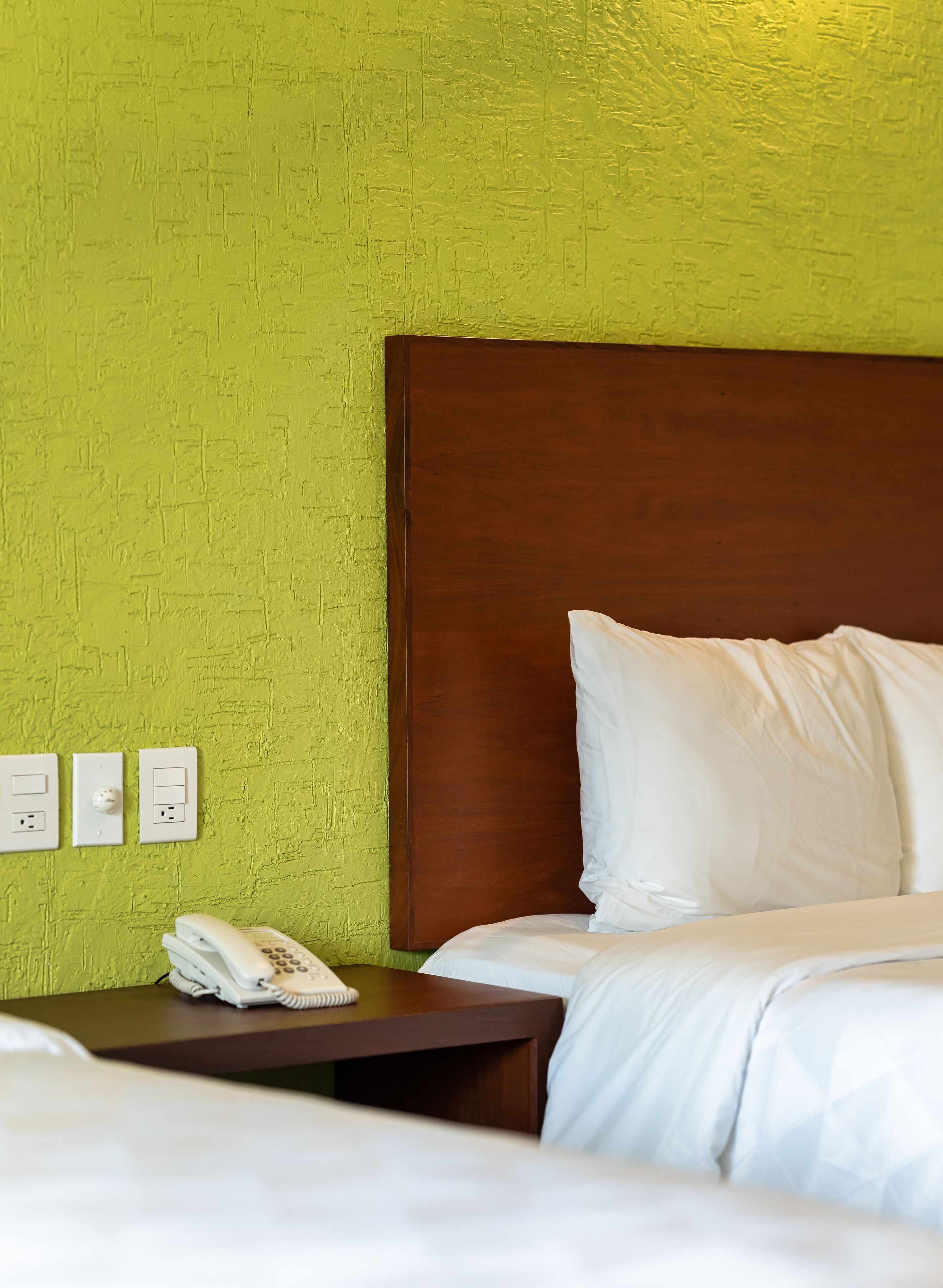 Ein Hotelzimmer mit einer grünen Wand mit Struktur, einem hölzernen Kopfteil, einem Nachttisch mit Telefon und einem Bett mit weißer Bettwäsche.