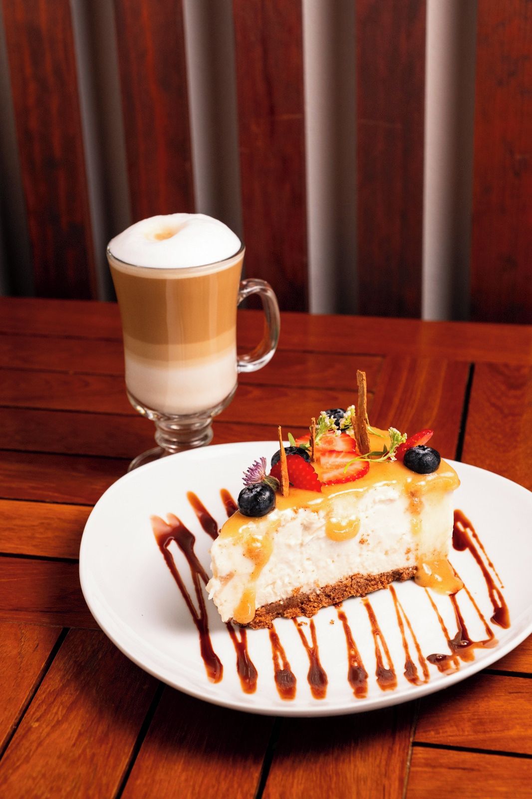 Un plato de postre con una porción de pastel de queso cubierto de fruta y salsa de caramelo, una taza de café en capas y una mesa de madera.