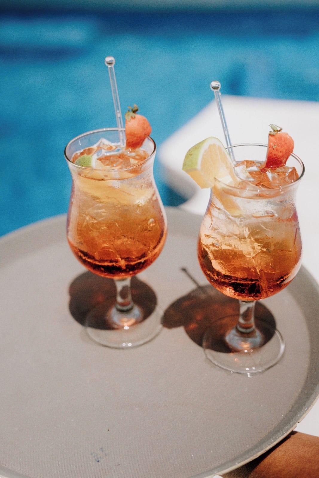 Two cocktails with garnishes, served in stemmed glasses, on a tray by a poolside.
