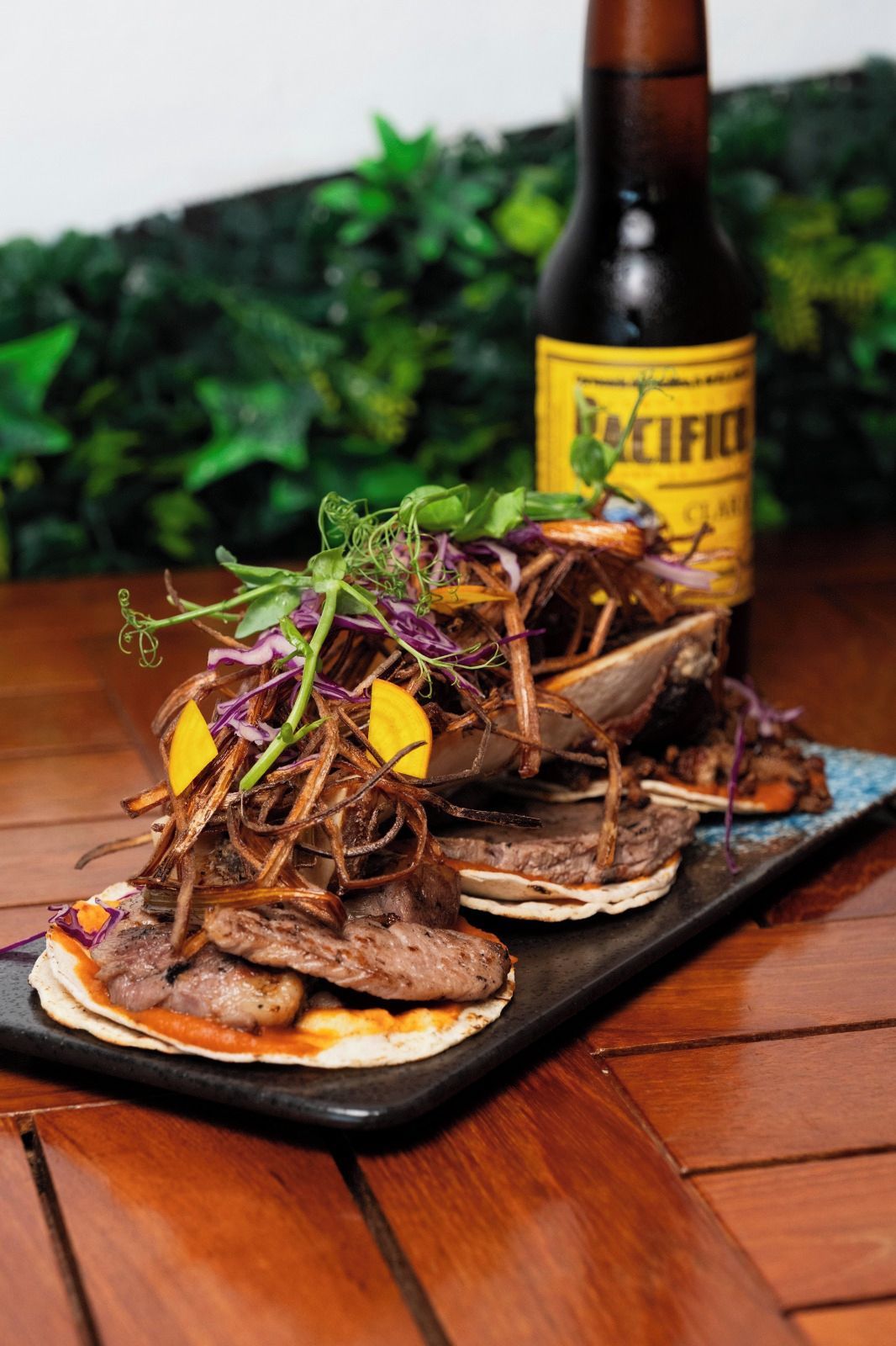 A wooden table with a black tray holding tacos topped with meat, vegetables, and a yellow sauce. A bottle labeled 'BICFIC' is in the background.