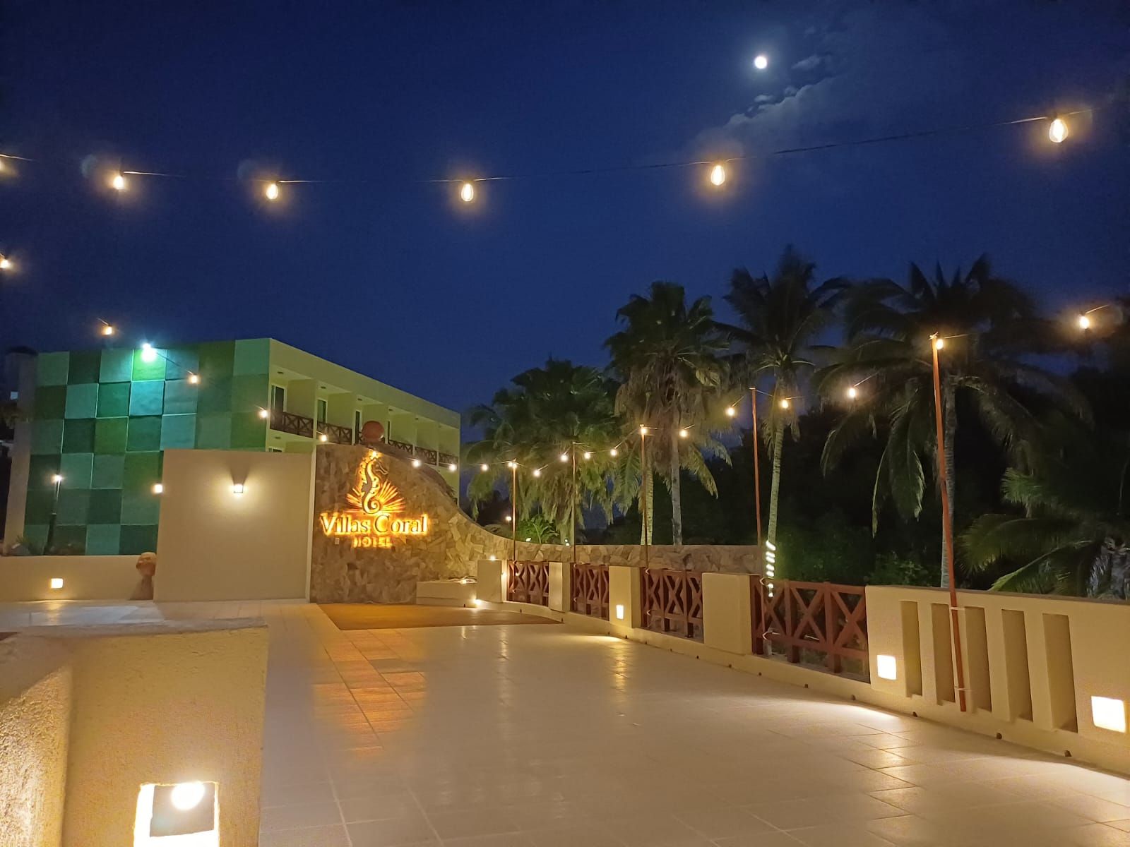 L’entrée d’un hôtel, avec une enseigne indiquant « Villas Coral Hotel », est bordée de palmiers et d’un chemin piétonnier dallé.