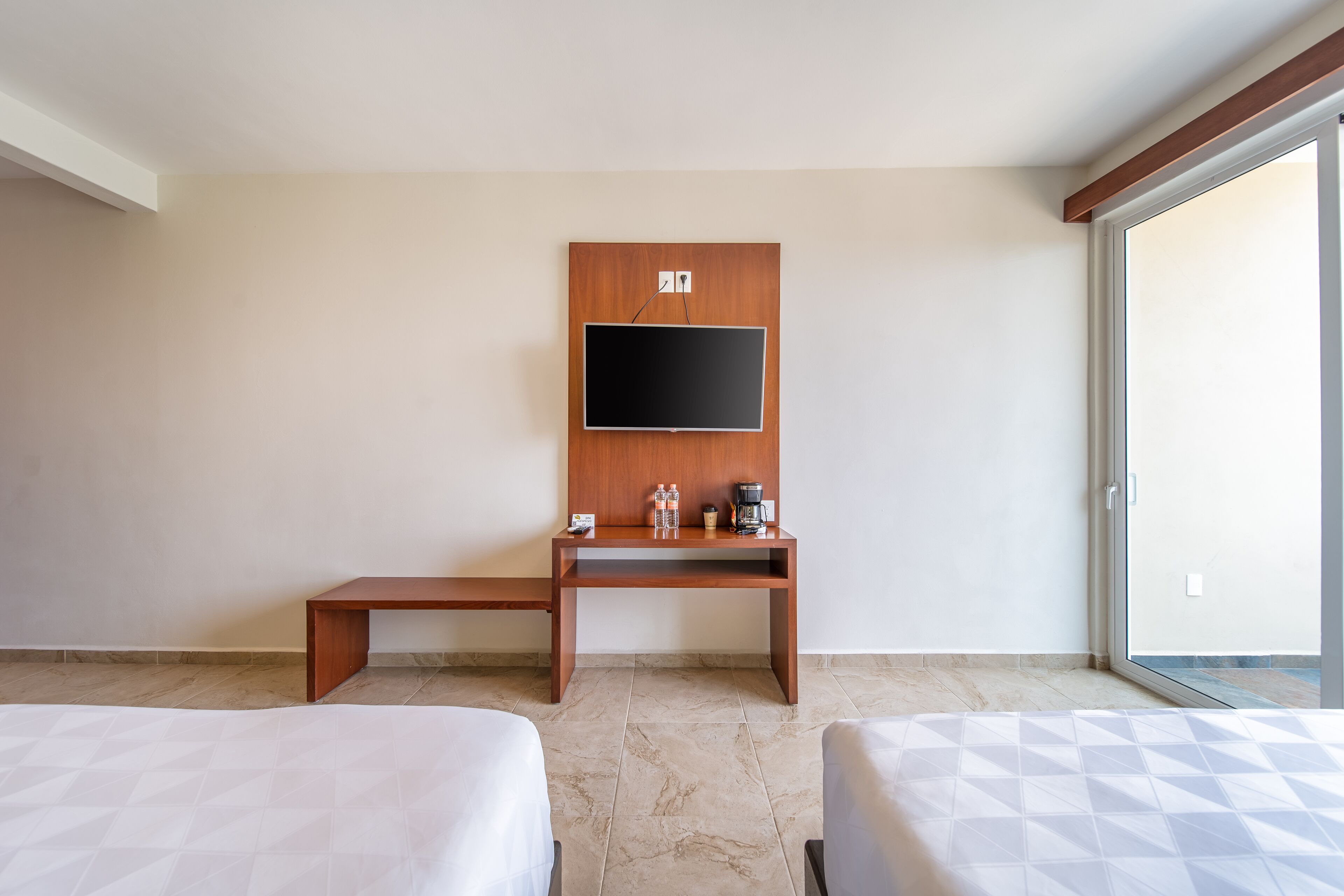 A hotel room with two beds, a wooden desk with a flat-screen TV, a coffee maker, and a bench.