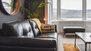 A modern living room with a leather sofa, a round mirror, a potted plant, and a view of the countryside through large windows. - 2 bedroom accommodation in Barmouth (Barmouth)