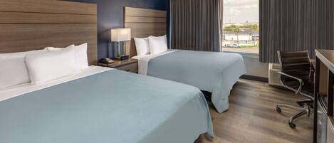 A hotel room with two beds, a desk, a chair, and a window with a view of the city.
