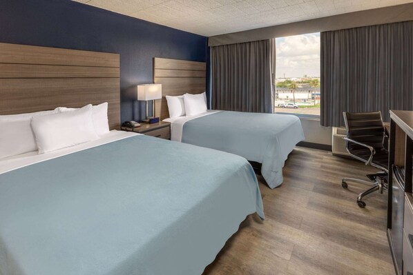 A hotel room with two beds, a desk, a chair, and a window with a view of the city.