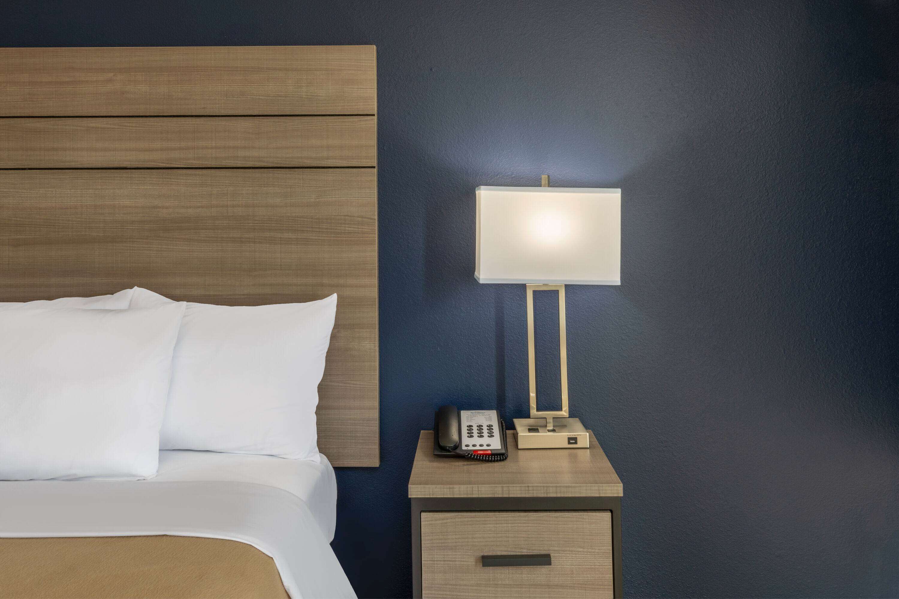 Uma cama bem arrumada com lençóis brancos, uma mesa de cabeceira com abajur, telefone e um pequeno móvel com gavetas.