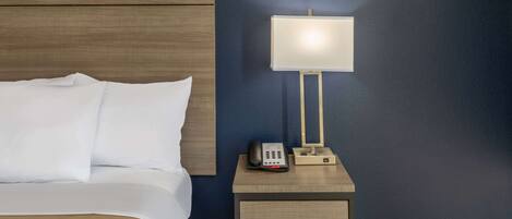 A neatly made bed with white linens, a bedside table with a lamp, a phone, and a small drawer unit.