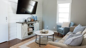 Una sala de estar con un televisor de pantalla plana montado en la pared, un mueble blanco para entretenimiento con estantes, una mesa de centro redonda, un sofá azul y una ventana con persianas.