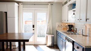 Una cocina con una mesa de madera, armarios blancos, lavavajillas y una ventana con cortinas.