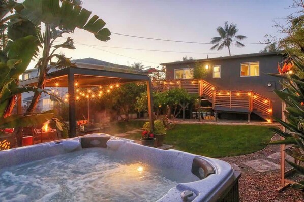 A hot tub in a backyard with a covered patio, a house with a balcony, and a grassy lawn.