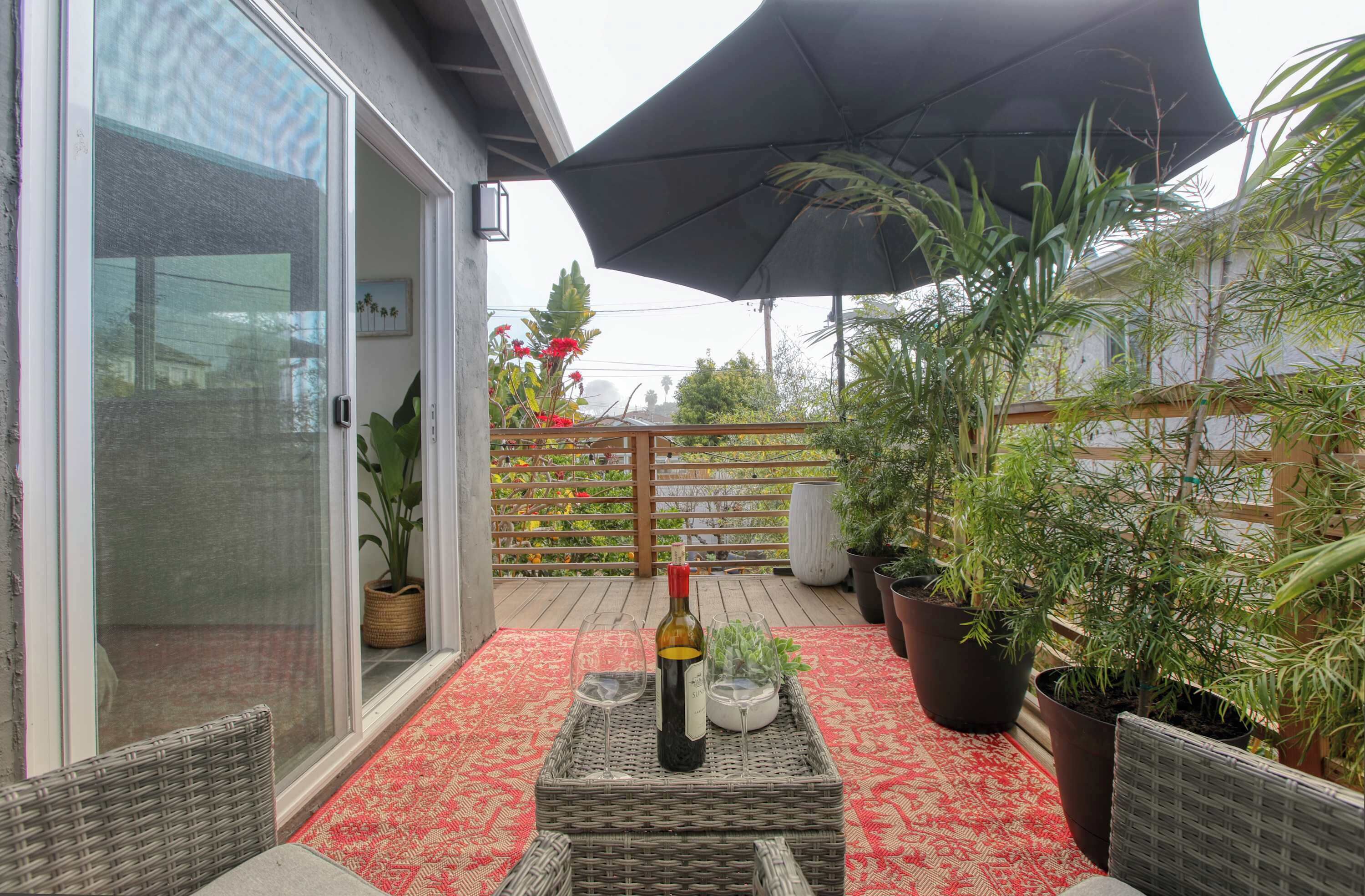 A covered patio with a red patterned rug, a wicker table with a bottle and glasses, potted plants, and a wooden fence.
