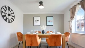 Ein Esszimmer mit einem Holztisch, sechs orangefarbenen Stühlen, einer schwarzen Uhr, zwei gerahmten Bildern und einem Fenster mit Vorhängen.
