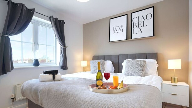 Ein Hotelzimmer mit einem Bett, Nachttischlampen, einem Fenster mit Vorhängen, einem Tablett mit Frühstücksgegenständen und zwei gerahmten Bildern an der Wand.