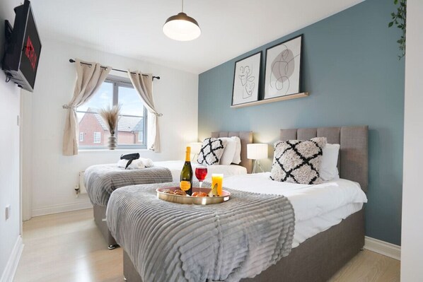 A bedroom with two beds, a window with curtains, a bedside table with a lamp, and artwork on the wall.