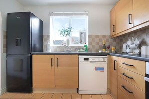 A kitchen with a black refrigerator, a dishwasher, wooden cabinets, a window with a plant, and a tiled backsplash.