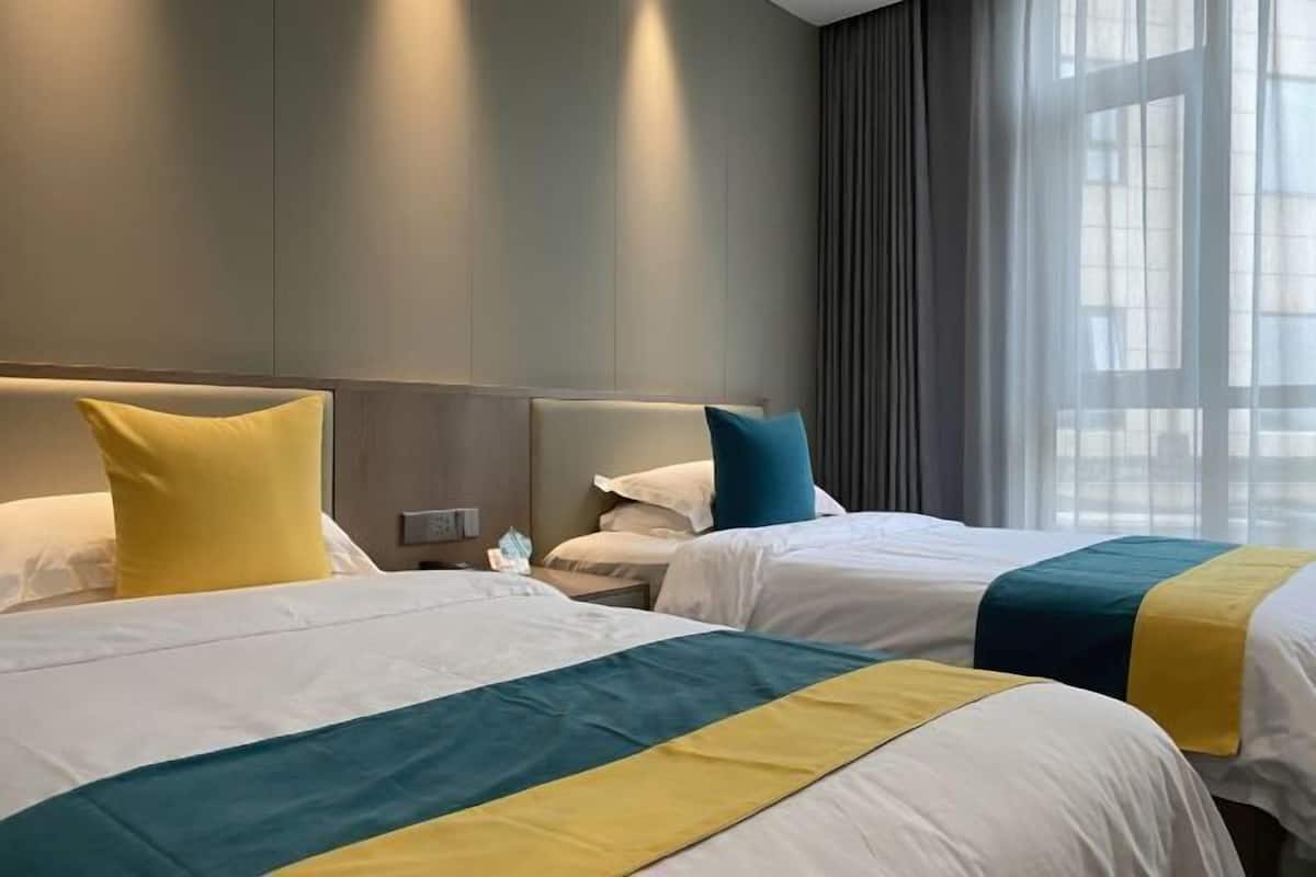 Quarto de hotel com duas camas, cada uma com colcha branca e azul-esverdeada, e uma mesa de cabeceira com abajur.