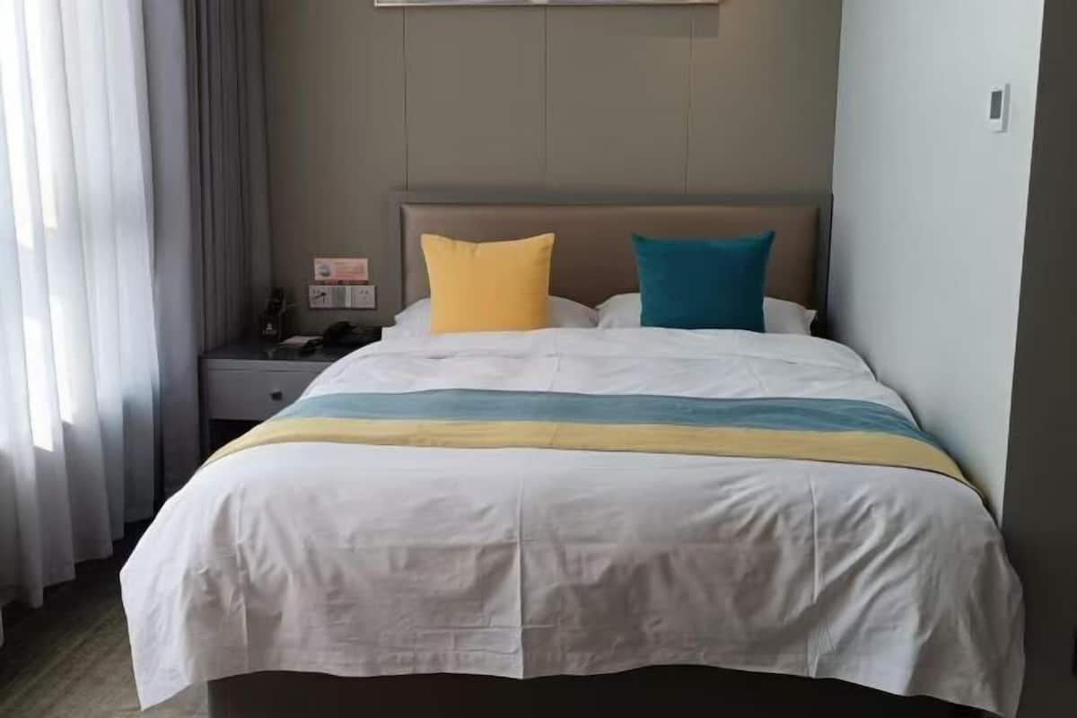 Quarto de hotel com cama, mesa de cabeceira, luminária e janela com cortinas.