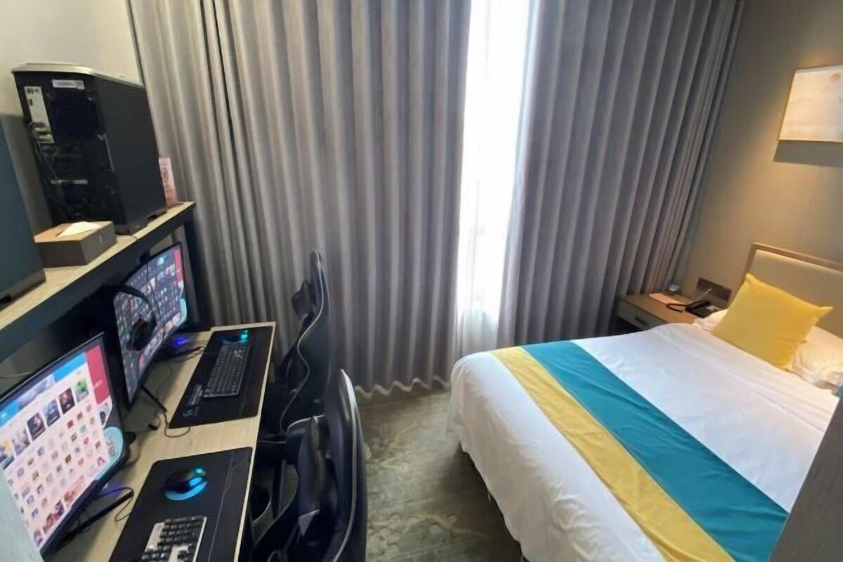 A hotel room with a bed, a desk with a computer, a chair, and a window with curtains.