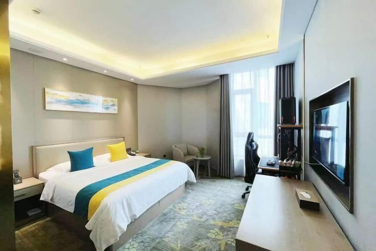 Quarto de hotel com cama, escrivaninha, cadeira, televisão e um quadro na parede.