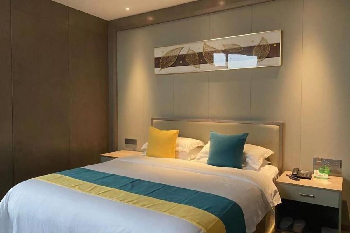 Quarto de hotel com cama, mesas de cabeceira, obra de arte na parede e mesa de apoio com telefone e uma pequena planta.
