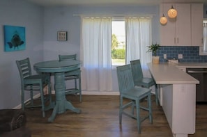Un coin repas avec une table ronde et quatre chaises, une cuisine avec des meubles blancs et un plan de travail bleu, et une fenêtre agrémentée de rideaux blancs.