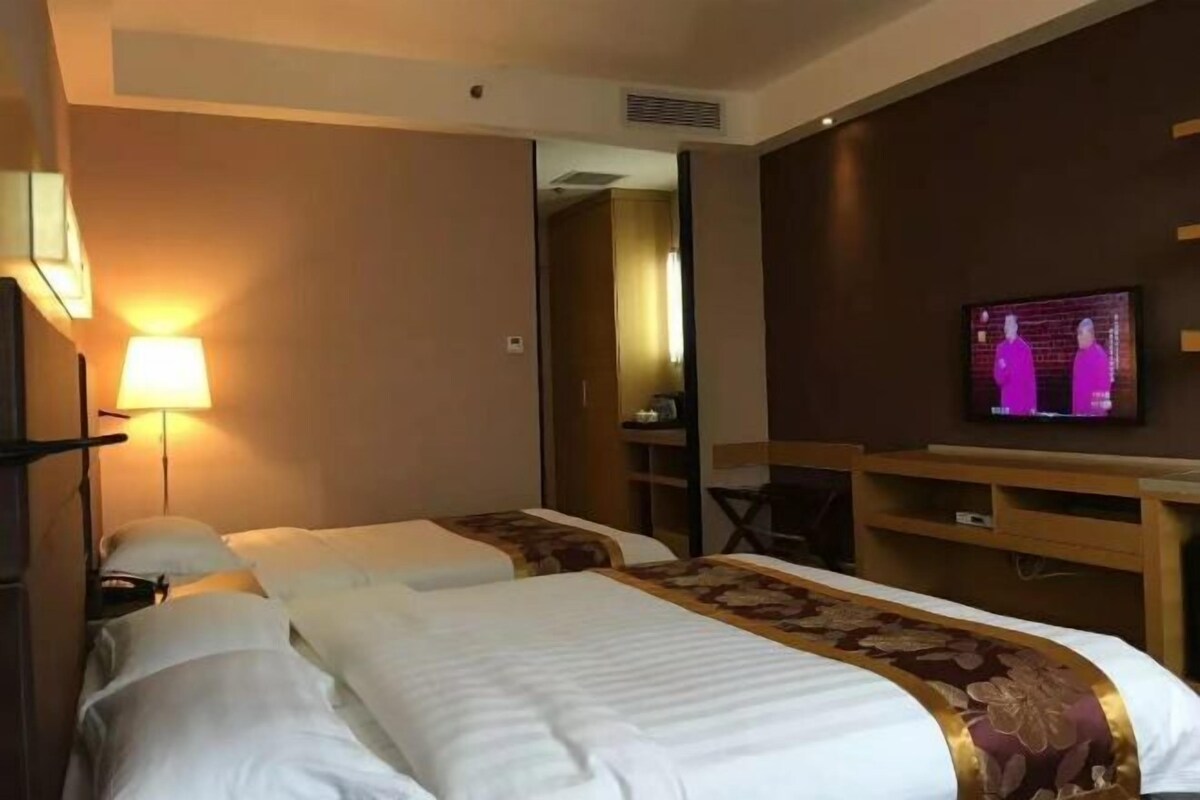 A hotel room with two beds, a TV mounted on the wall, a desk, and a lamp.