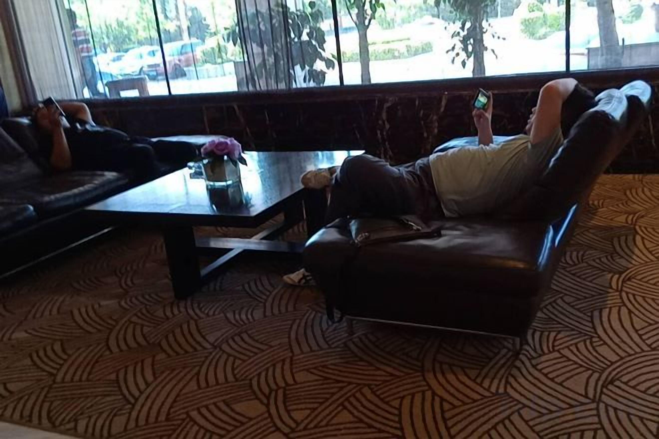 Una persona recostada en un sofá de cuero en una habitación con alfombra estampada, una mesita de café y una ventana con vistas a coches aparcados y árboles.