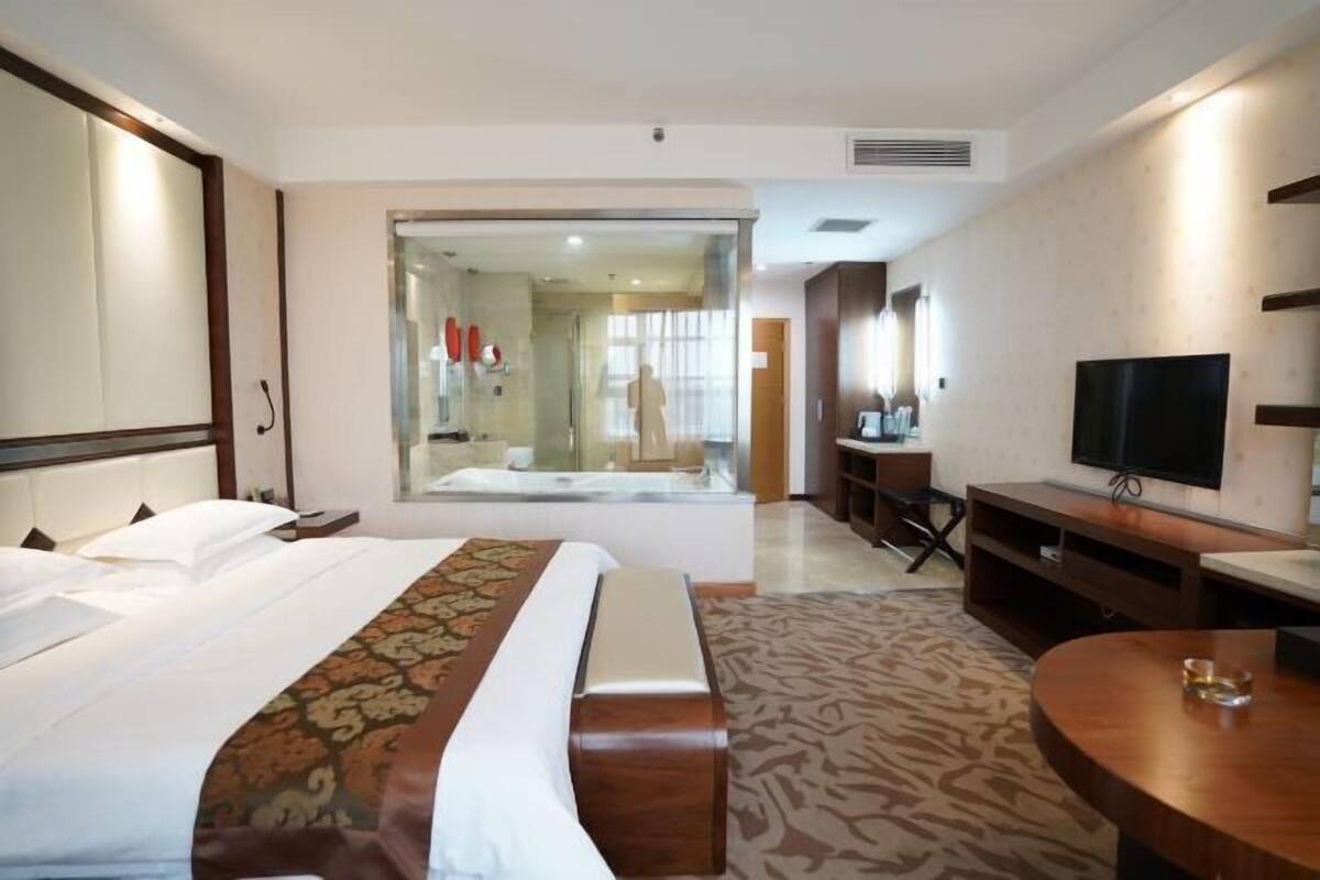 A hotel room with a bed, a TV, a desk, and a glass-enclosed bathroom.