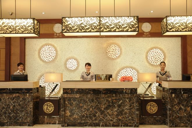 Zona de recepción de hotel con tres recepcionistas sentadas tras mostradores, cada una con una lámpara y un arreglo floral. La decoración incluye adornos circulares en la pared y un techo con motivos.