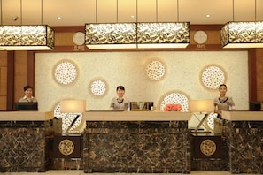 A hotel reception area with three receptionists seated behind counters, each with a lamp and a floral arrangement. The decor includes circular wall decorations and a patterned ceiling.