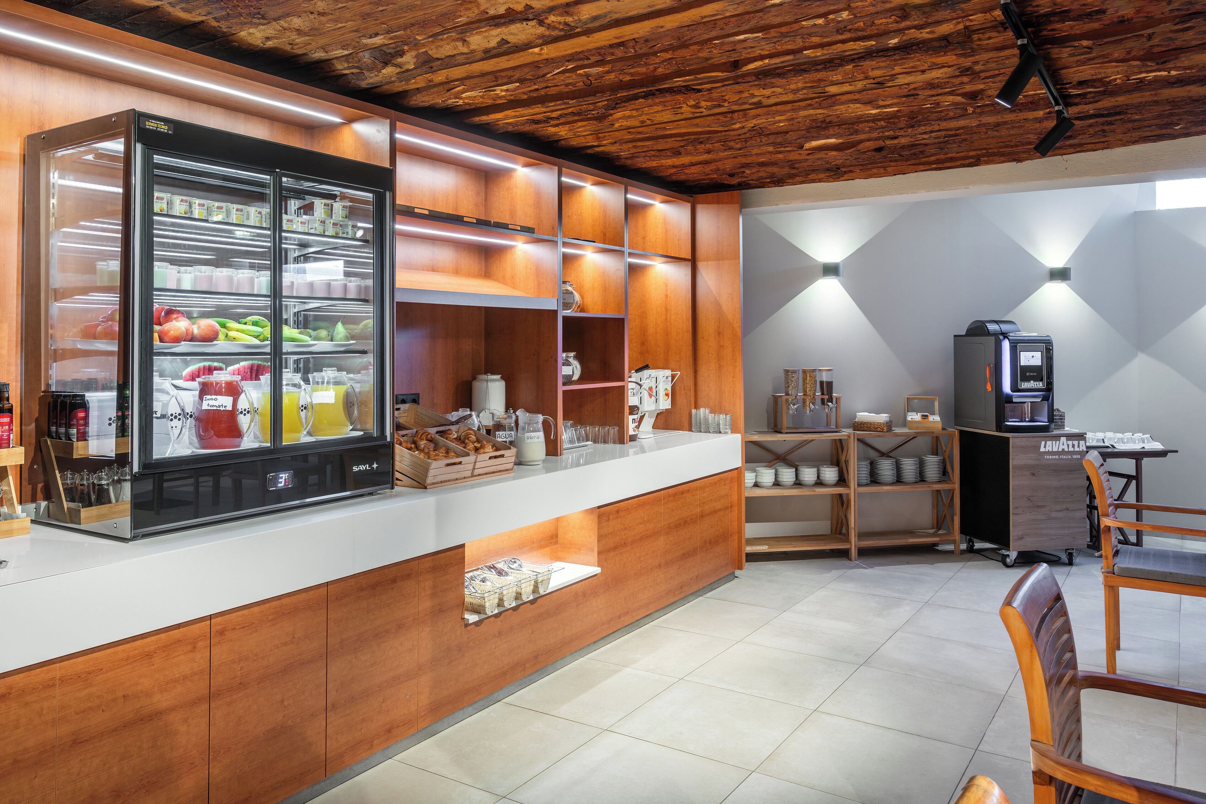 A modern kitchen area with a refrigerated display case, wooden cabinets, and a coffee machine.