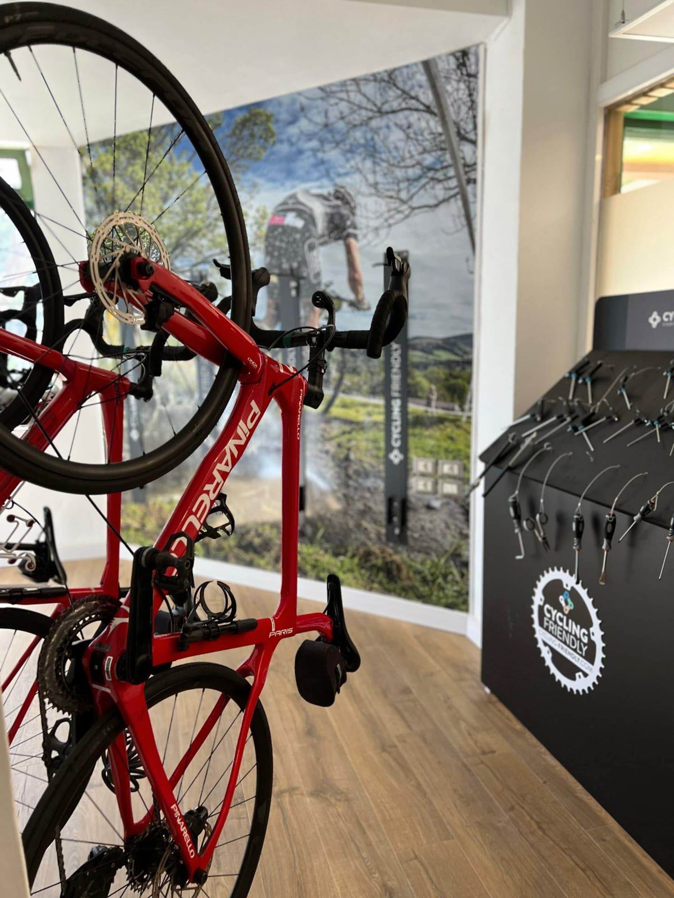 Una bicicleta roja con el nombre de marca 'Peyrele' en el cuadro, ubicada en un espacio interior con un cartel de fondo y una exhibición de accesorios de ciclismo a la derecha.