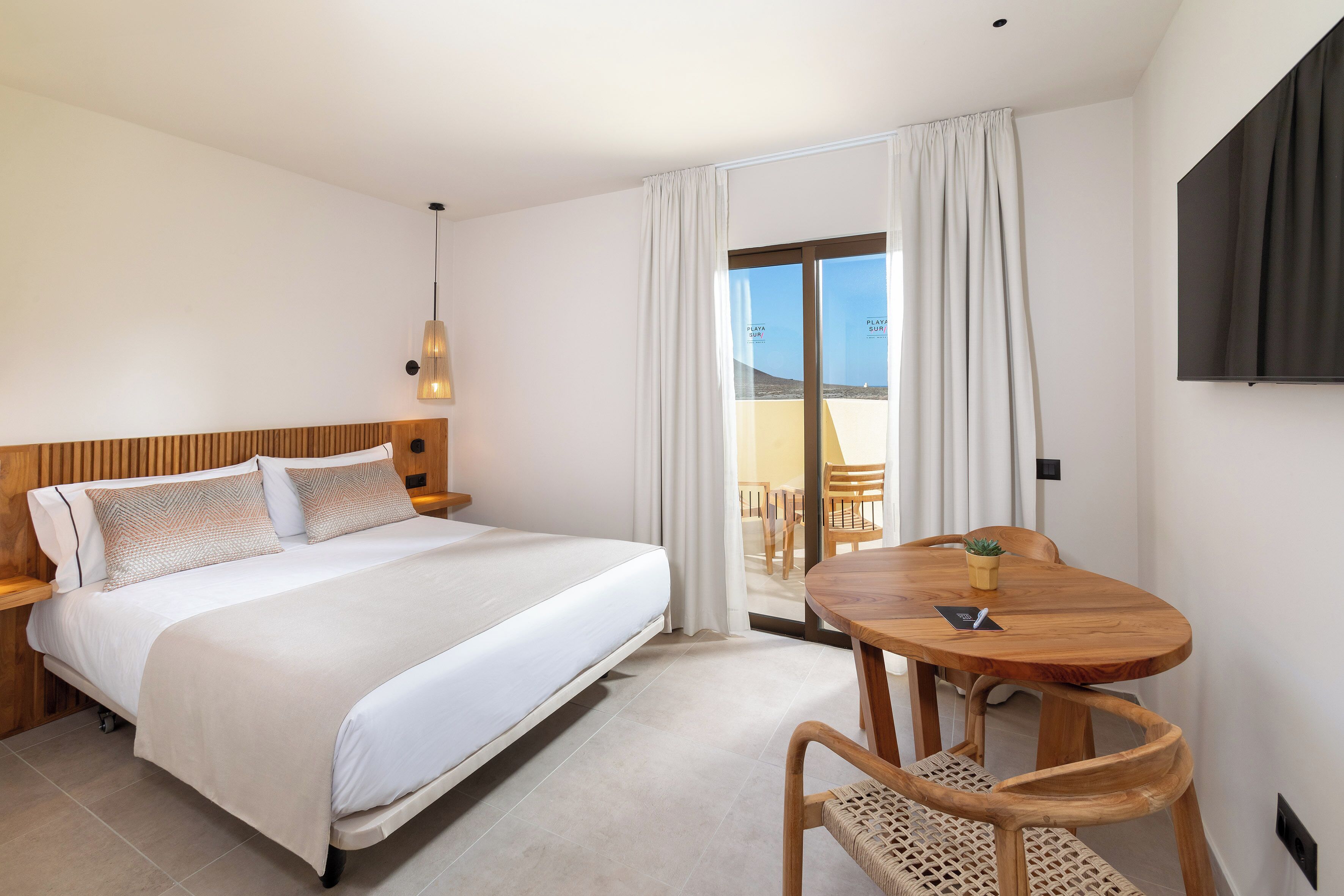 Une chambre d’hôtel comprenant un lit, une tête de lit en bois, une petite table en bois avec une chaise, une plante en pot et une porte-fenêtre coulissante donnant sur un espace extérieur.