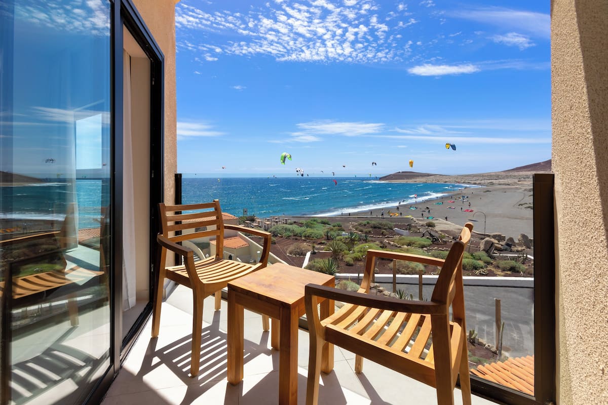 A balcony with wooden furniture overlooking a beach with people and kites.