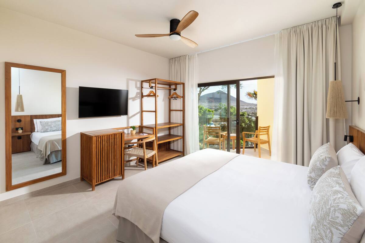 A modern hotel room with a large bed, a wooden desk, a television, and a balcony with a view of the outdoors.
