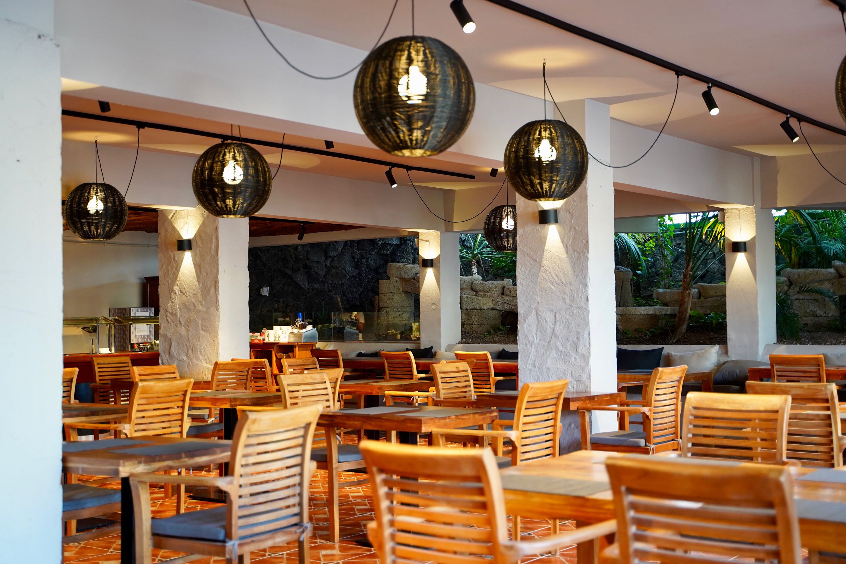 Un espace de restauration intérieur avec des tables et des chaises en bois, de grandes lampes sphériques décoratives et une vue sur un cadre naturel grâce à de grandes fenêtres.