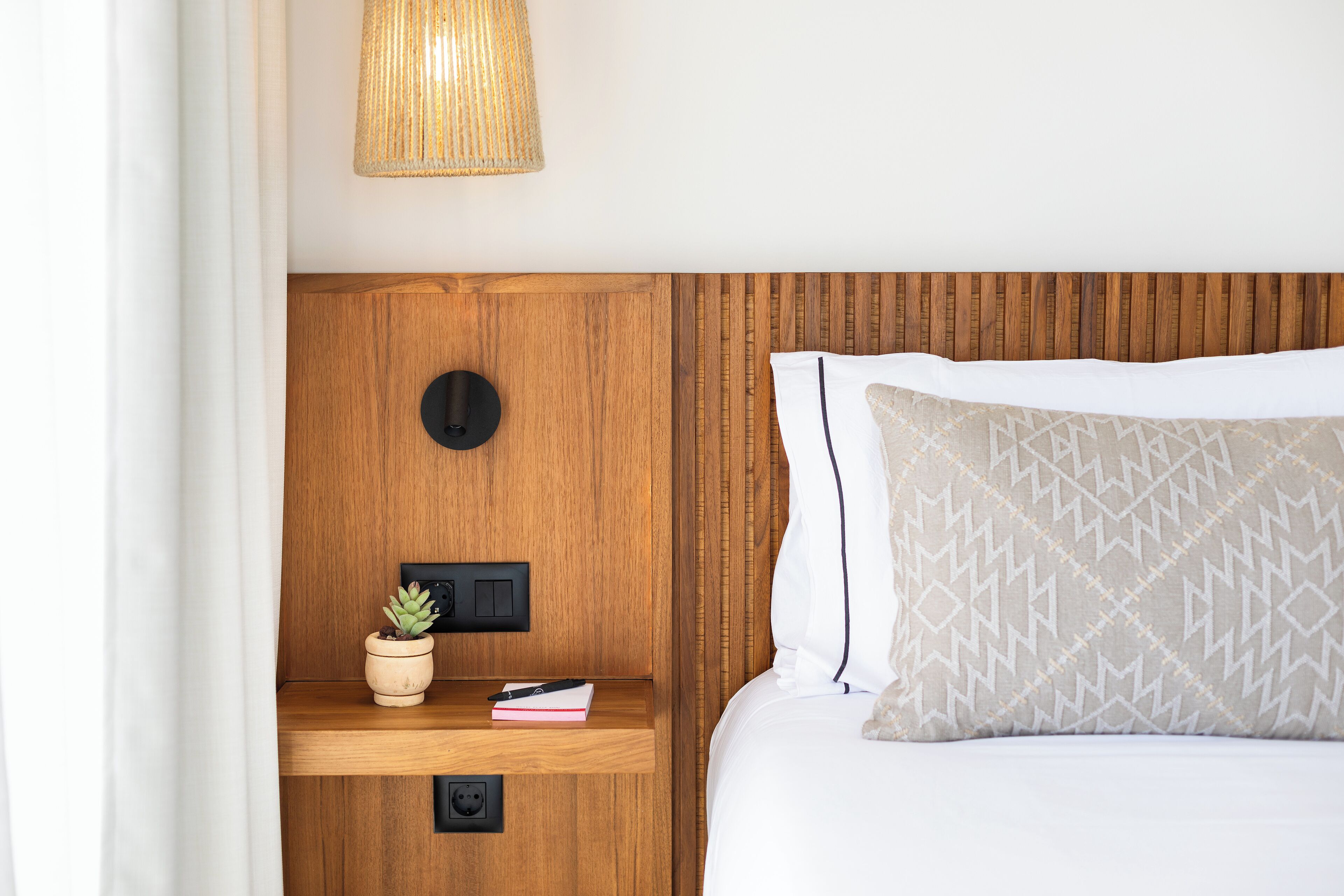 A neatly made bed with a patterned pillow, a bedside lamp, a small plant, and a nightstand with a book and remote control.