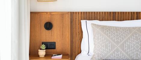 A neatly made bed with a patterned pillow, a bedside lamp, a small plant, and a nightstand with a book and remote control.