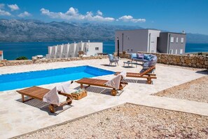 Un espace piscine doté de chaises longues, d’un mur en pierre et offrant une vue sur la mer et les montagnes.