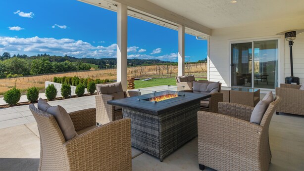 An outdoor patio with wicker furniture, a glass-top table with a fire feature, and a scenic view of a vineyard.