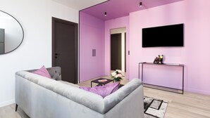 Ein modernes Wohnzimmer mit grauem Sofa, einem kleinen Tisch mit Blumenvase, einem Flachbildfernseher an einer lilafarbenen Wand und einer Tür zu einem anderen Raum.