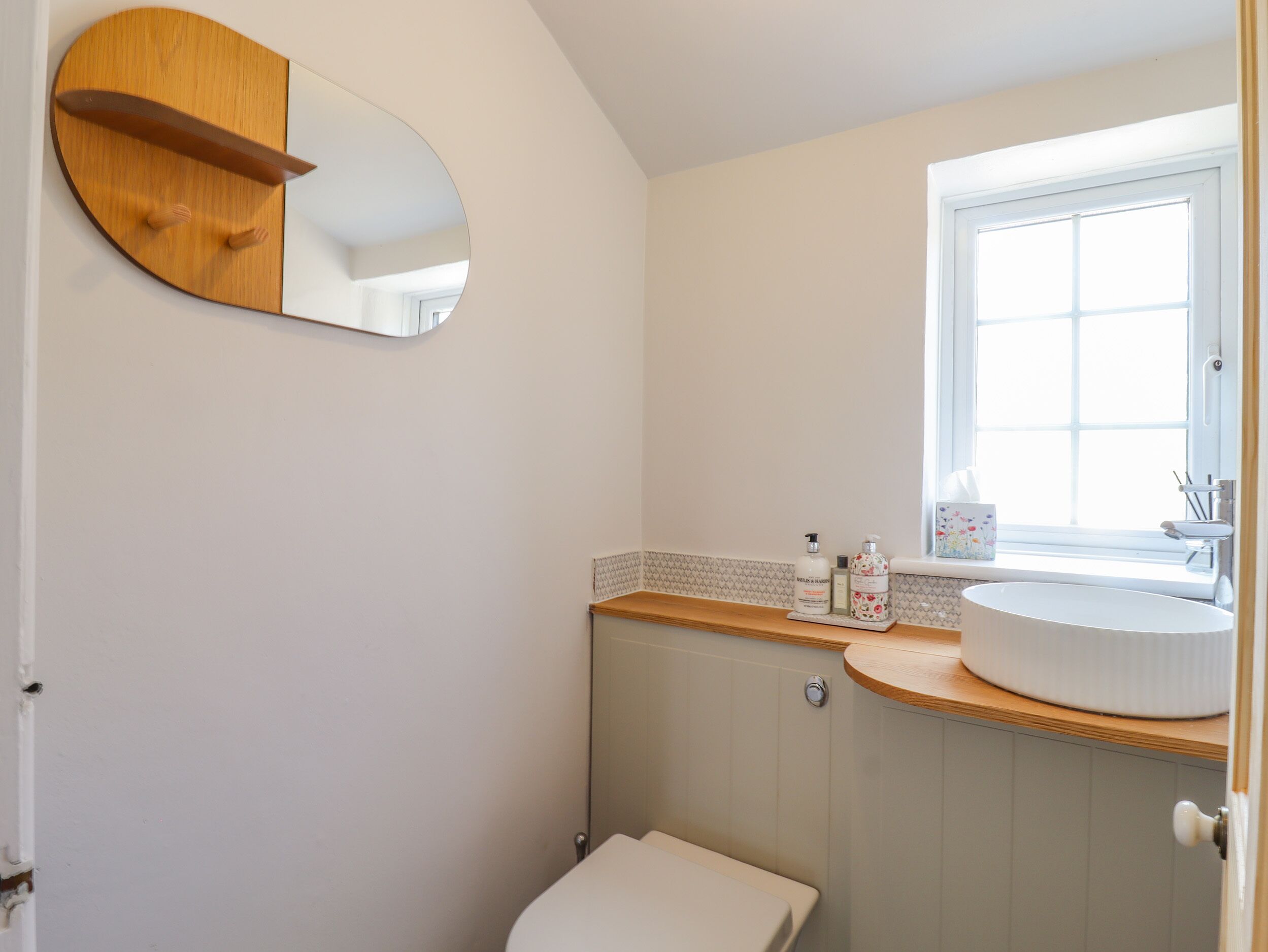 Un baño con espejo redondo, repisa de madera, inodoro blanco y ventana.