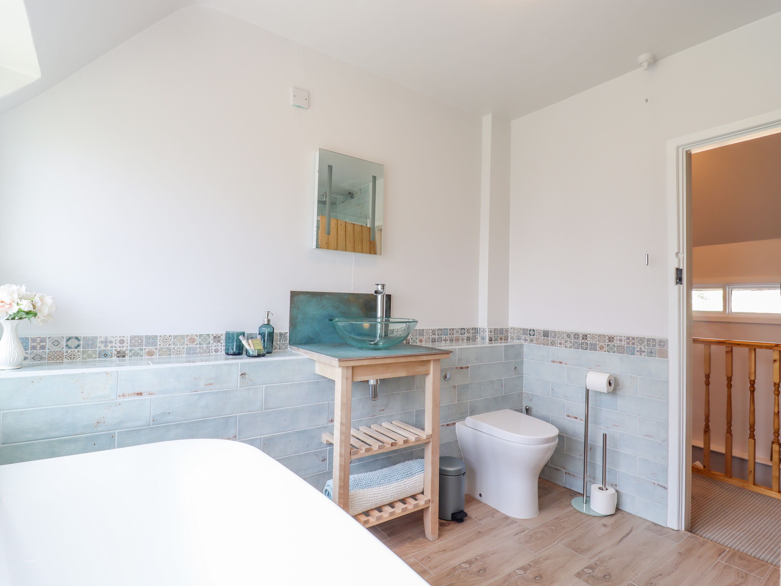 A bathroom with a glass bowl sink, a toilet, a wooden shelf, a trash bin, and a mirror on the wall.