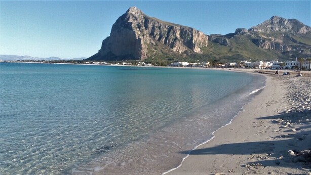 Ein Strand mit klarem Wasser, felsigem Ufer und einem Berg im Hintergrund.