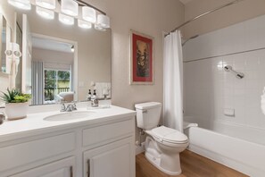 Een badkamer met een wit wastafelmeubel, toilet en bad, een potplant en een lijst met een foto aan de muur.